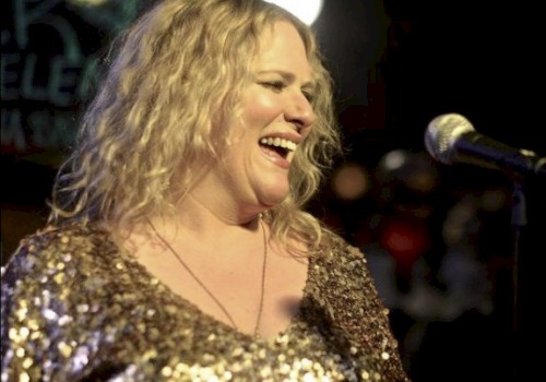 A person is smiling and standing in front of a microphone, wearing a sparkly outfit, with blurred lights in the background.