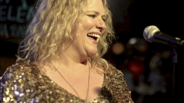A person is smiling and standing in front of a microphone, wearing a sparkly outfit, with blurred lights in the background.