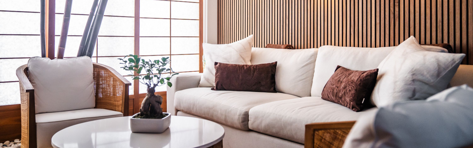 A modern living room with white furniture, brown cushions, wooden panel walls, warm lighting, and a small table with a plant in the center.
