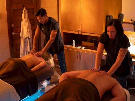 Two people receiving massages in a dimly lit room, with therapists working on them. There's a cozy, relaxing atmosphere.