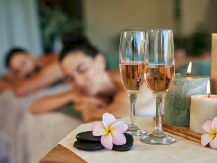 A couple are relaxing face-down on massage tables, while two glasses of rosé, candles, and flowers create a calming spa atmosphere.