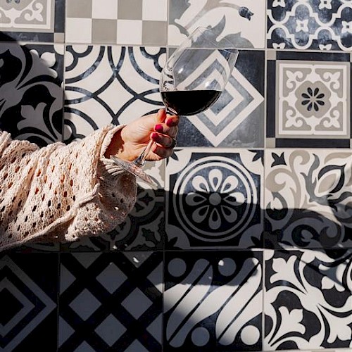 A hand holding a wine glass is in front of a wall with assorted black and white patterned tiles, creating a stylish backdrop.