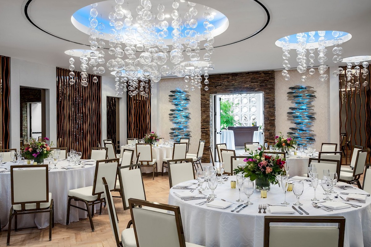 An elegantly decorated banquet hall with round tables, floral centerpieces, and modern chandeliers, ready for an event.