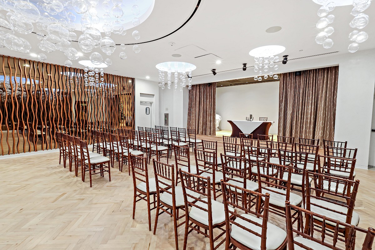 An elegant, modern event space with rows of wooden chairs, decorative hanging lights, and a small table at the front, ready for a gathering.