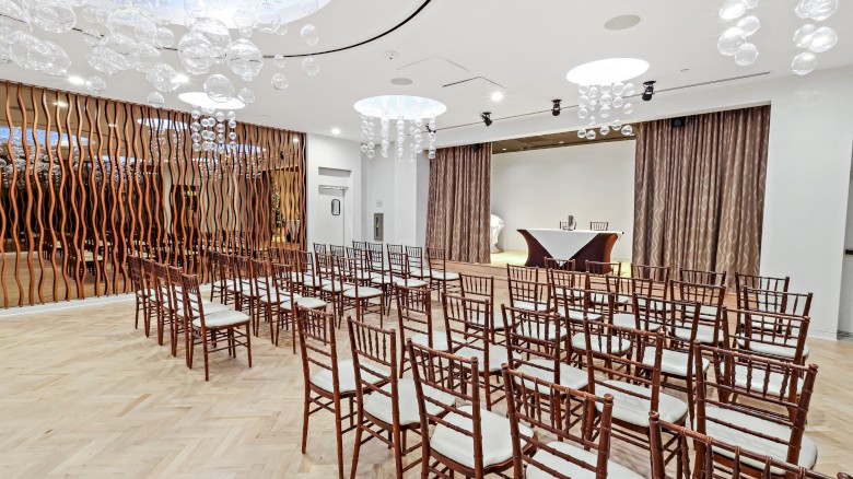 An elegant, modern event space with rows of wooden chairs, decorative hanging lights, and a small table at the front, ready for a gathering.