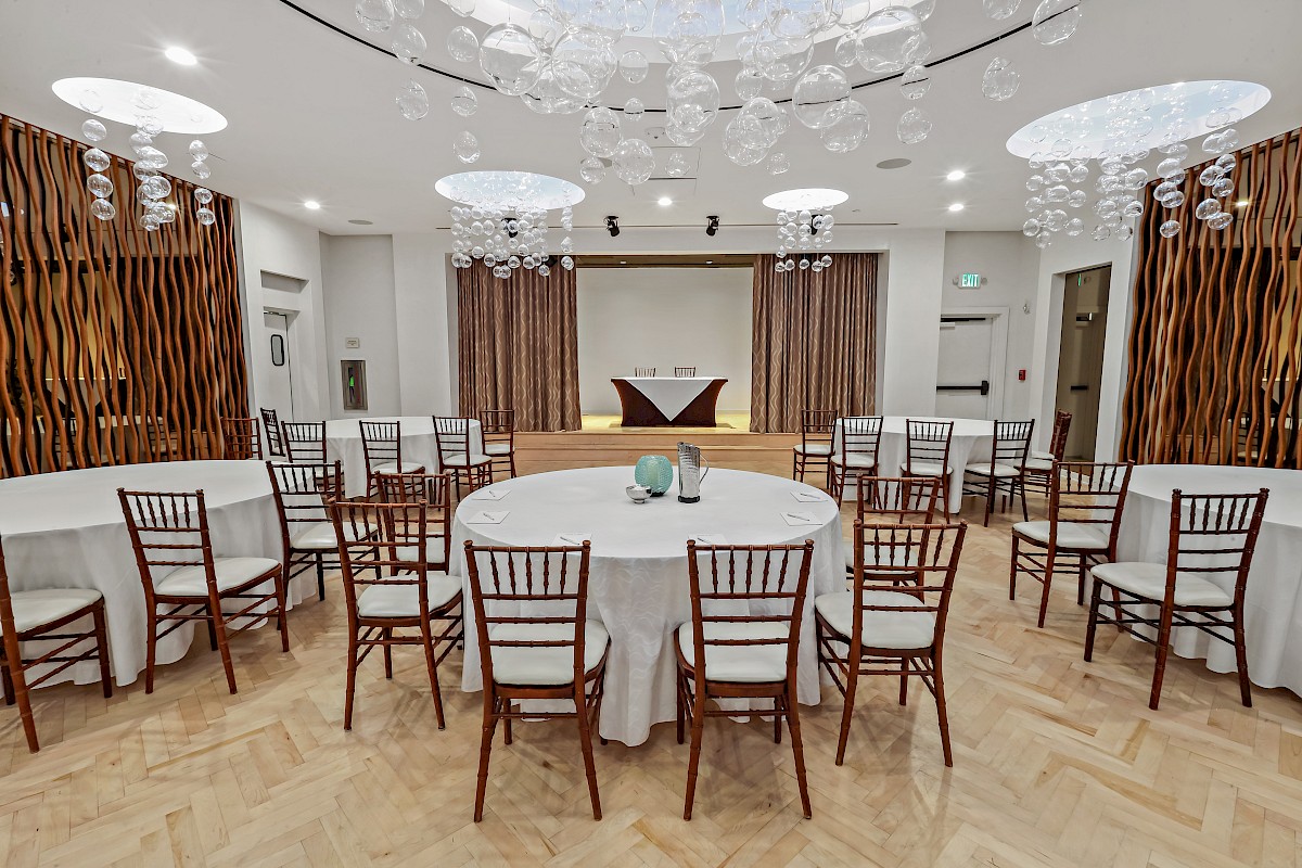 The image shows an event space with round tables, wooden chairs, and modern decor, set beneath artistic ceiling lights.