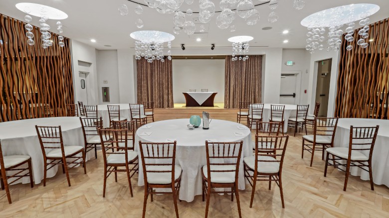 The image shows an event space with round tables, wooden chairs, and modern decor, set beneath artistic ceiling lights.