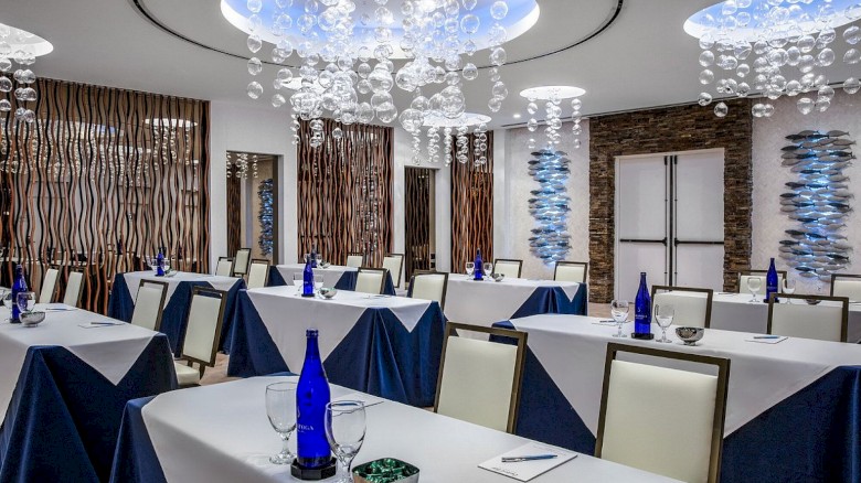 A modern meeting room with tables set with blue bottles, glasses, and notepads, featuring decorative ceilings and wood panel accents.