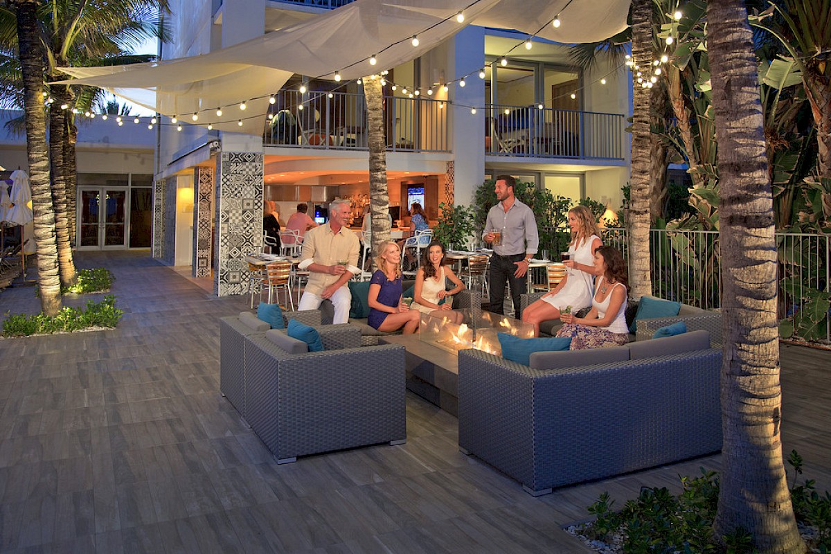 A group of people are socializing around a fire pit on a patio with string lights and palm trees, giving a warm, festive atmosphere.