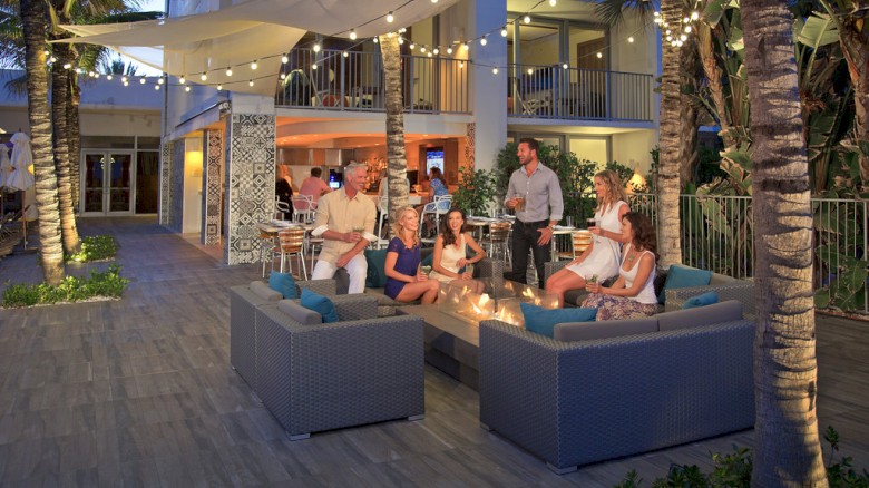 A group of people are socializing around a fire pit on a patio with string lights and palm trees, giving a warm, festive atmosphere.