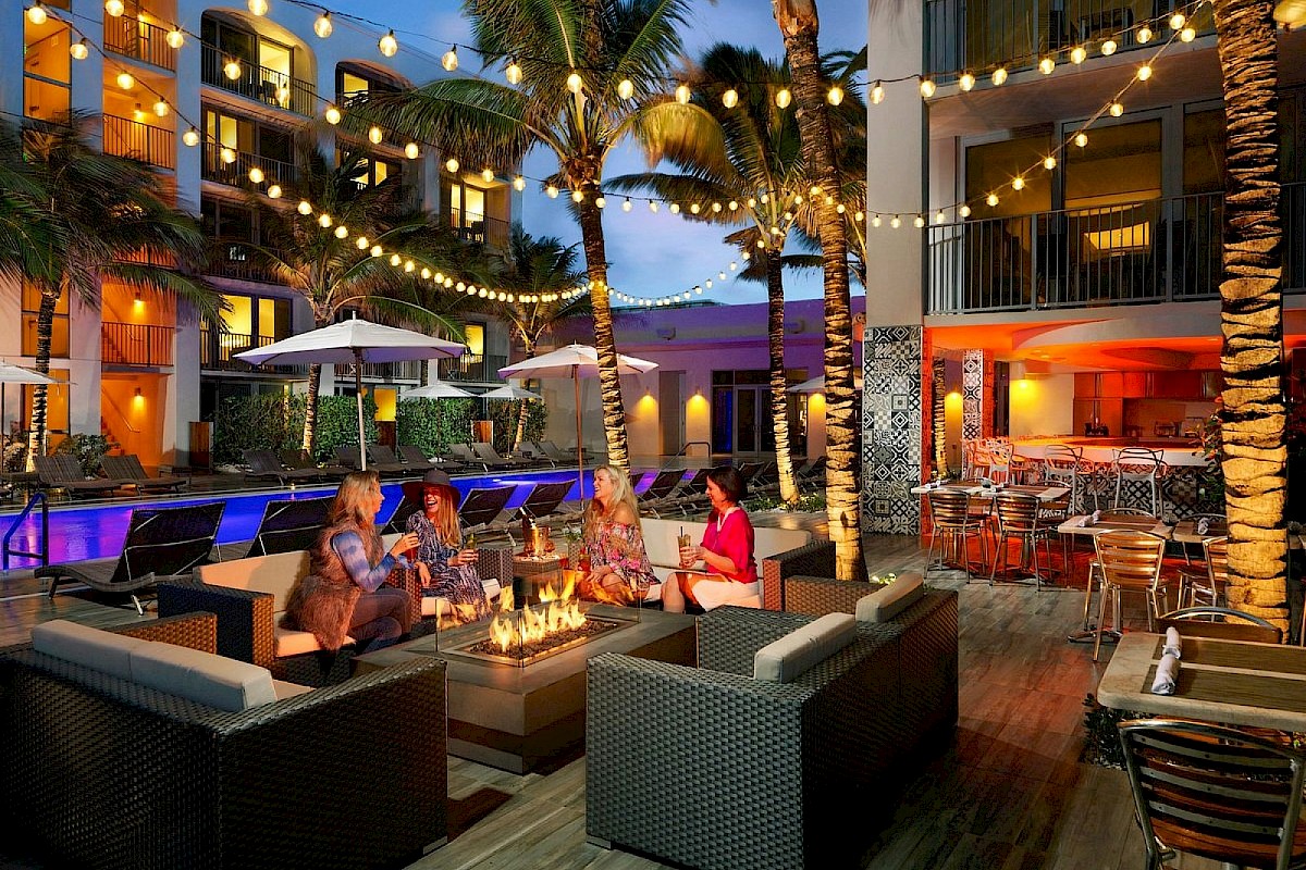 Outdoor seating area by a pool with string lights, palm trees, and people around a fire pit. Cozy and inviting atmosphere.