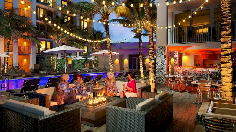 Outdoor seating area by a pool with string lights, palm trees, and people around a fire pit. Cozy and inviting atmosphere.