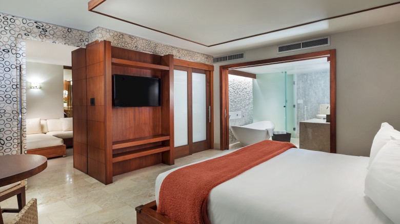 The image shows a modern hotel room with a large bed, TV, wooden furnishings, and an open bathroom featuring a bathtub and glass shower.
