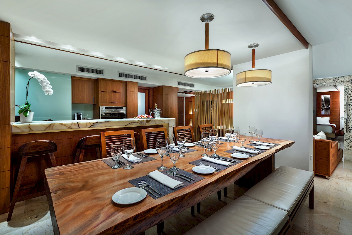 A modern dining area with a wooden table, set with plates and glasses. A kitchen in the background features wooden cabinets and an orchid.