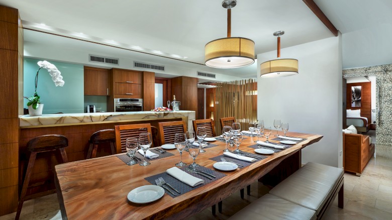 A modern dining area with a wooden table, set with plates and glasses. A kitchen in the background features wooden cabinets and an orchid.