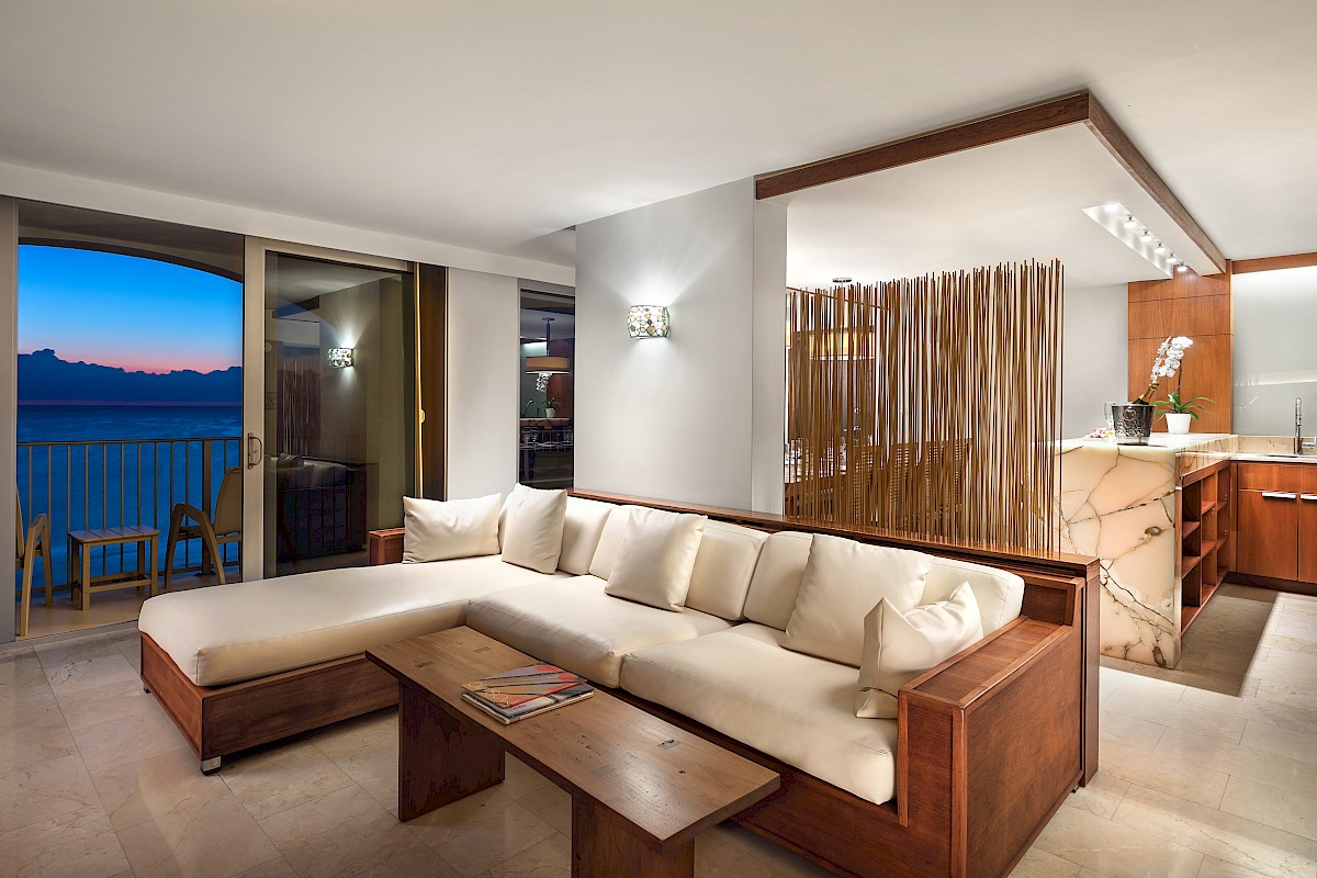 A modern living room features a white sectional sofa, wooden accents, and a view of the ocean through sliding doors at sunset.