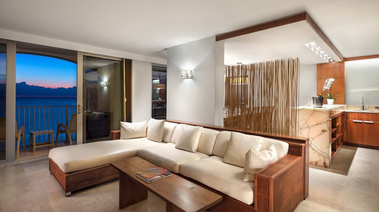 A modern living room features a white sectional sofa, wooden accents, and a view of the ocean through sliding doors at sunset.