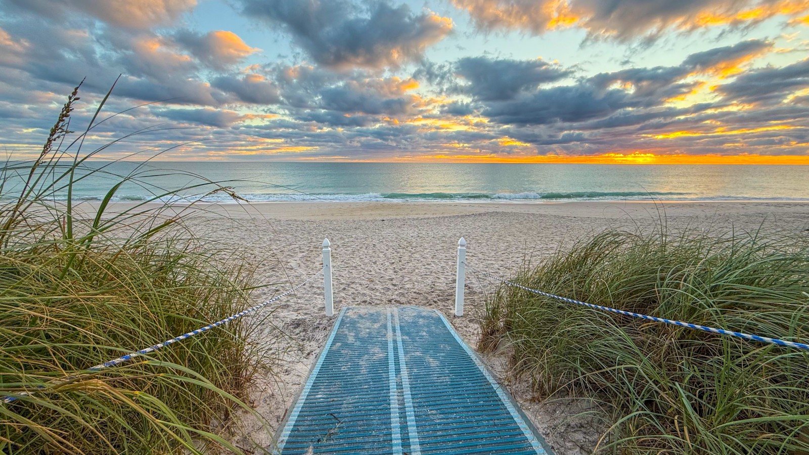 A sunrise over a peaceful beach with a pathway through dunes leading to the ocean and colorful clouds in the sky.