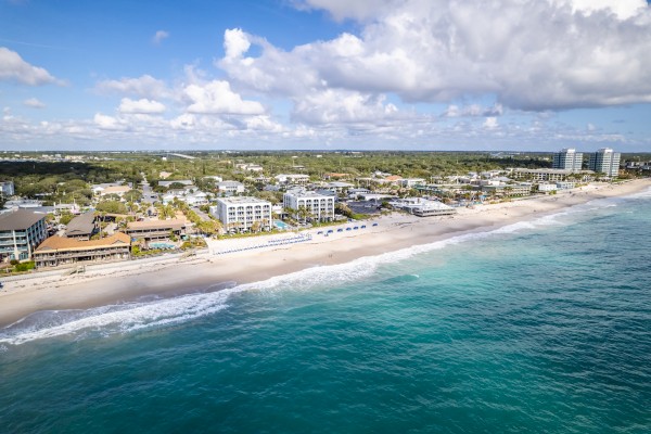 A scenic beach with turquoise water, sandy shore, modern beachfront buildings, and a partly cloudy sky.