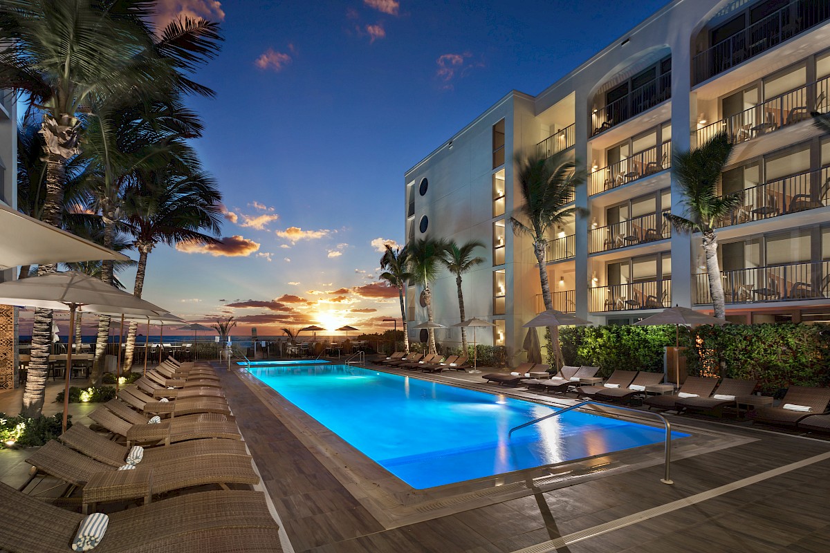 A modern hotel pool area at sunset with palm trees, lounge chairs, and a view of the ocean in the background.