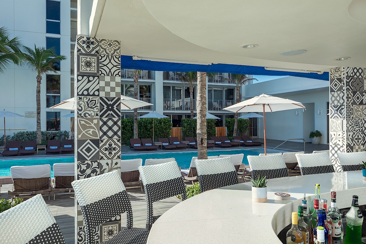 Poolside bar with patterned columns, stools, and liquor bottles. Lounge chairs and umbrellas by the pool, under clear skies.