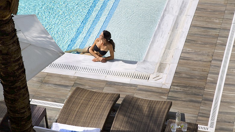 A person relaxes at the edge of a pool with two lounge chairs and towels nearby, under an umbrella.