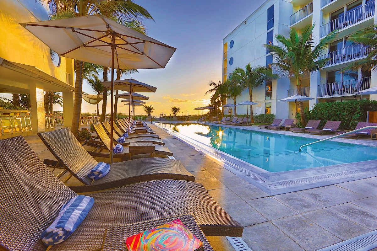 A luxurious poolside scene with lounge chairs, umbrellas, and palm trees at sunset, adjacent to a modern building exterior.