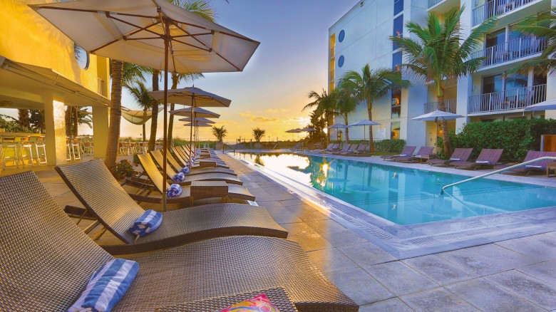 A luxurious poolside scene with lounge chairs, umbrellas, and palm trees at sunset, adjacent to a modern building exterior.