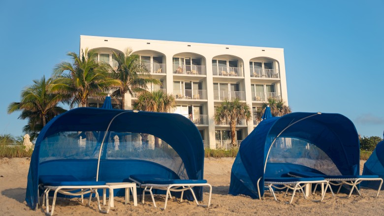 Beach scene with blue cabanas and lounge chairs on sand, in front of a white building with balconies and palm trees under a clear sky.