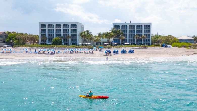 A person in a kayak is on the bright blue ocean near a sandy beach. Buildings and beach chairs line the shore under a cloudy sky.