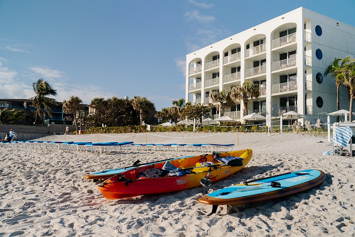 A beach scene with kayaks and paddleboards on the sand, near a white multi-story building with balconies and surrounded by palm trees.