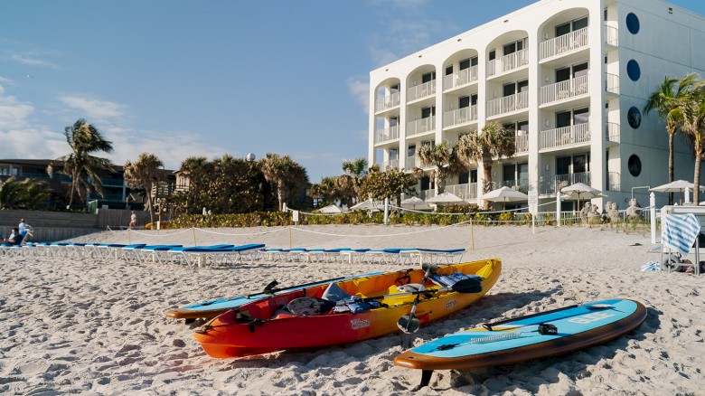 A beach scene with kayaks and paddleboards on the sand, near a white multi-story building with balconies and surrounded by palm trees.