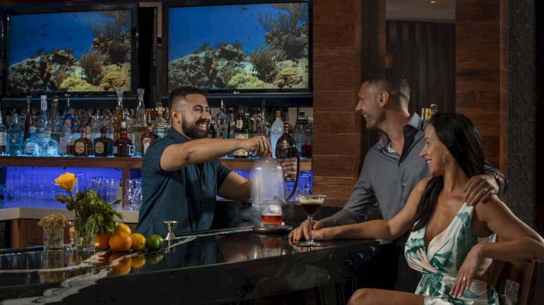 A bartender serves drinks to a couple at a bar, surrounded by fruits and a scenic marine backdrop on screens.