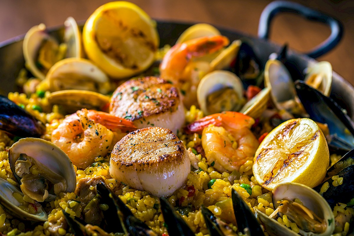 A pan of paella featuring seafood like scallops, shrimp, clams, and mussels on saffron rice, garnished with lemon wedges.