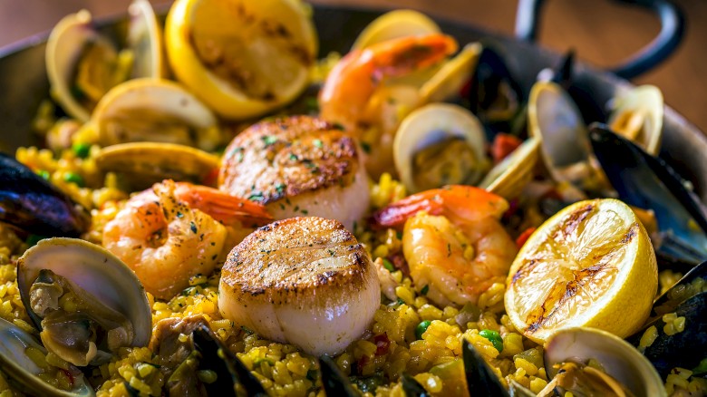 A pan of paella featuring seafood like scallops, shrimp, clams, and mussels on saffron rice, garnished with lemon wedges.