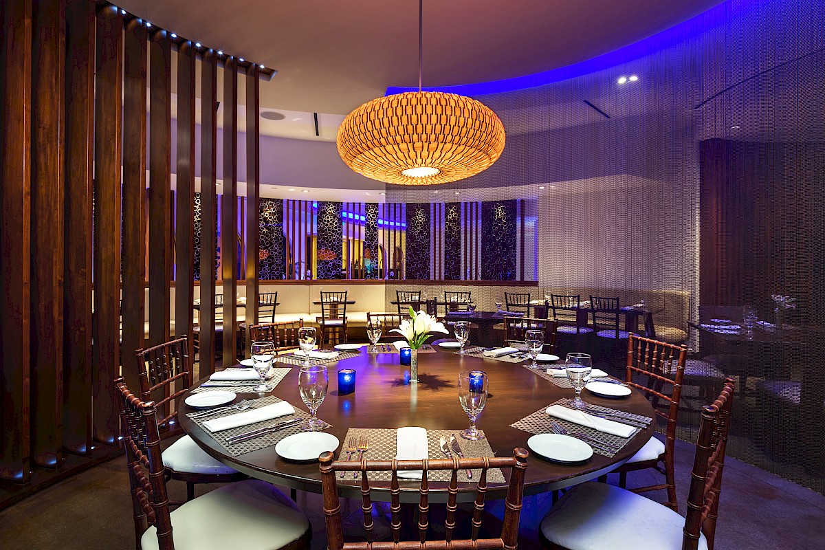 A modern dining room with a large round table set for eight, featuring elegant lighting and decor in a stylish ambiance.