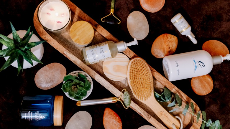 The image shows a flatlay of spa items like a candle, brush, jade roller, skincare bottles, and plants on a textured background.