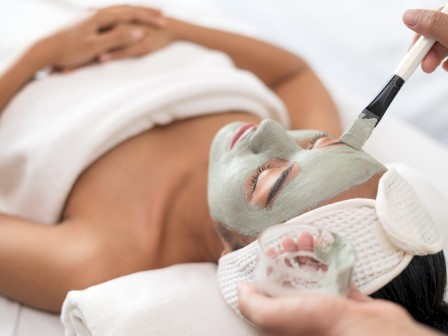 A person is receiving a facial treatment with a green face mask being applied using a brush, wearing a headband and towel on a spa bed.