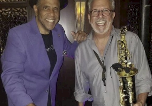 Two smiling men, one holding bongos, the other a saxophone, in a music-themed setting, wearing casual attire.