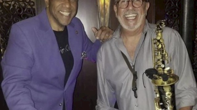 Two smiling men, one holding bongos, the other a saxophone, in a music-themed setting, wearing casual attire.