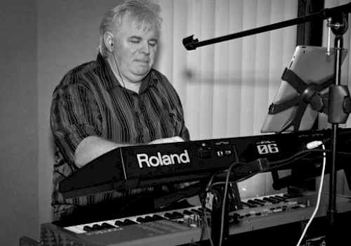 A person is playing a Roland keyboard, with headphones on, sheet music on a stand, and a microphone nearby.