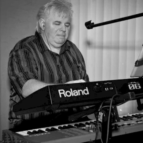 A person is playing a Roland keyboard, with headphones on, sheet music on a stand, and a microphone nearby.