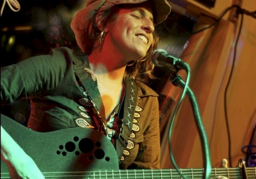 A person is playing guitar and singing into a microphone, wearing a hat and smiling, in a warmly lit setting.
