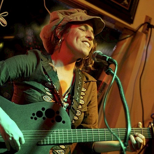 A person is playing guitar and singing into a microphone, wearing a hat and smiling, in a warmly lit setting.