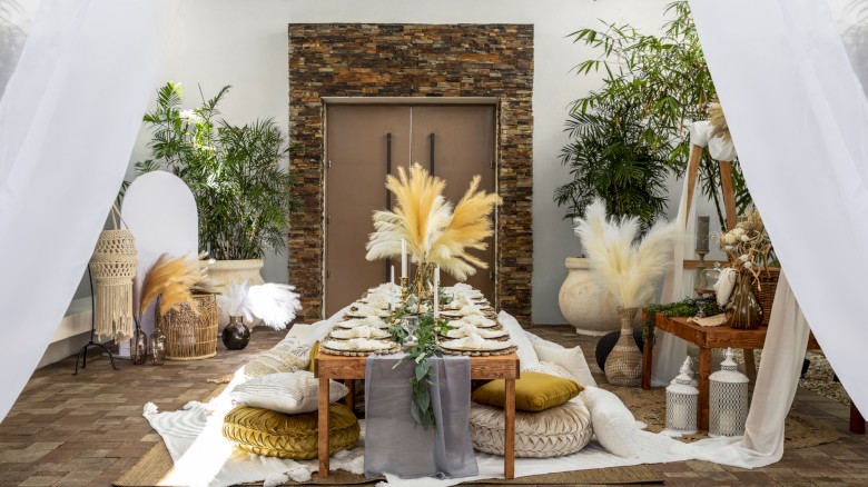 Elegantly set outdoor dining area with rustic decor, pampas grass centerpieces, pillows, and lush plants, under sheer white drapes.