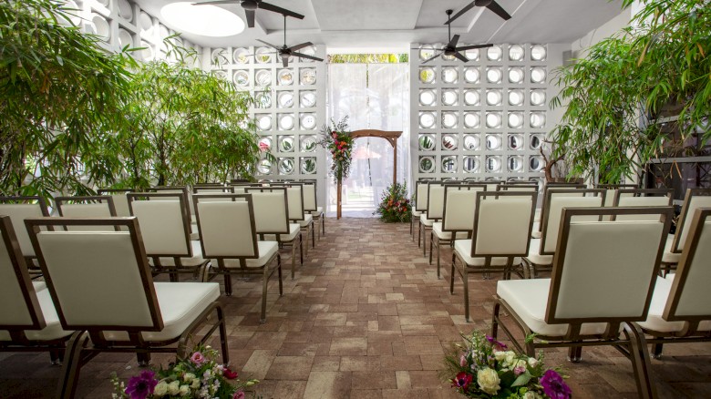 The image shows a wedding ceremony setup with chairs, floral arrangements, and green plants in a bright, open space.