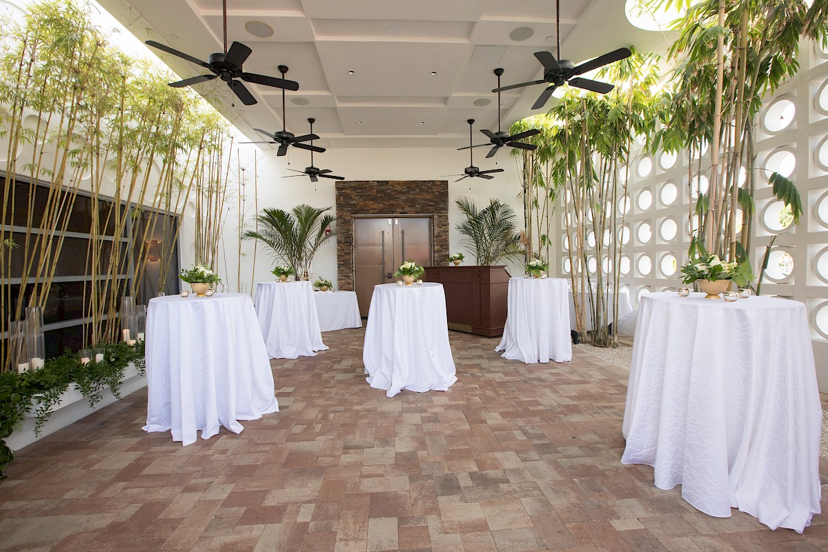 An elegant room with tall tables covered in white cloth, surrounded by bamboo, plants, and ceiling fans in a modern setting.