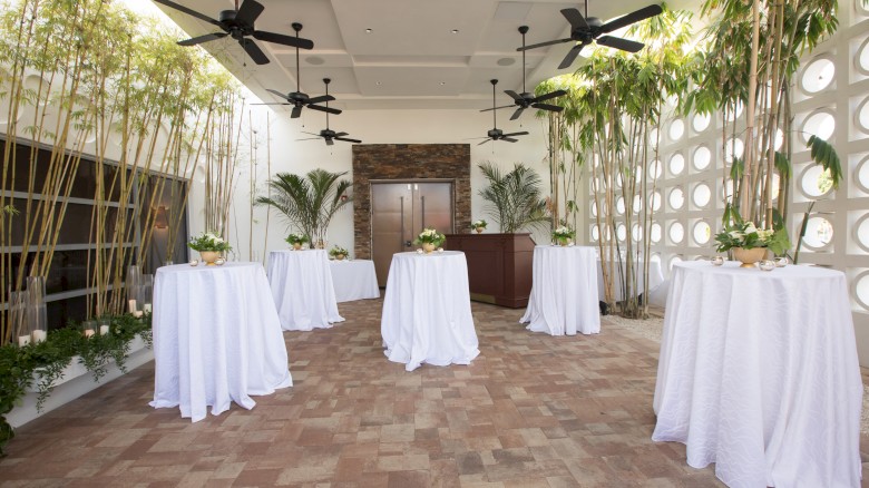 An elegant room with tall tables covered in white cloth, surrounded by bamboo, plants, and ceiling fans in a modern setting.