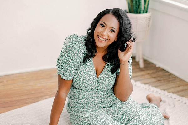 A person sits on a blanket with vintage cameras and a magazine, near a plant in a well-lit room, smiling at the camera.