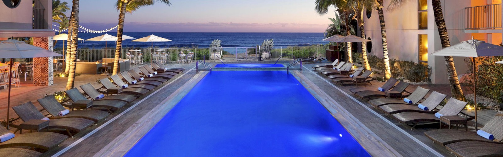 An illuminated swimming pool surrounded by lounge chairs and palm trees at a resort, with an oceanside view at sunset.